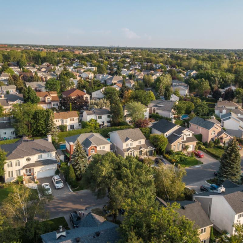 Suburban neighborhood seen from above, showing residential rooftops surrounded by trees on a clear day.